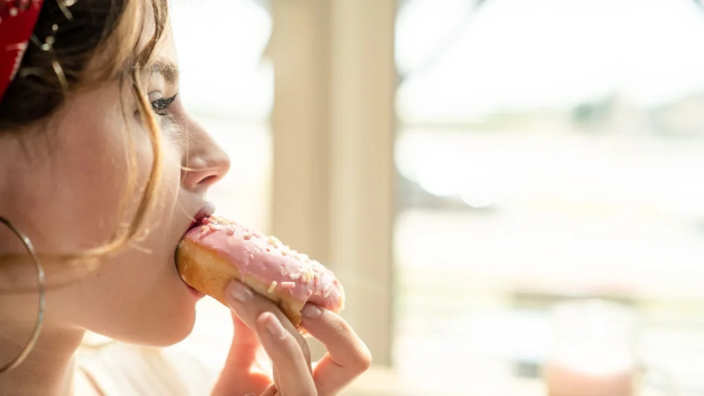 Pembe kaplamalı bir donut yiyen kadın; aniden bastıran şeker krizini ve duygusal açlığı temsil eden görsel.